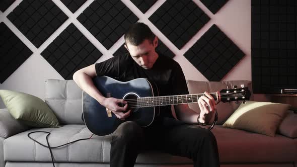 Young Man Playing Acoustic Guitar While Sitting on Sofa in Home Recording Studio alt