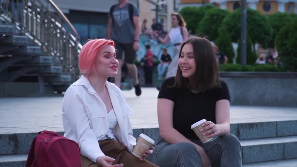 Two Young Women Talking and Laughing Sitting on the Granite Stairs and Drinking Coffee alt