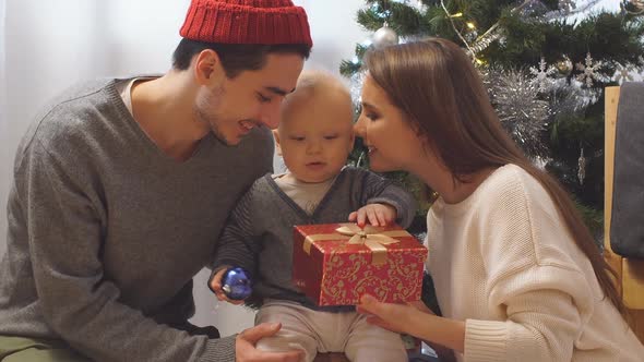 Happy Family and Baby with Christmas Gift Near the Christmas Tree at Home. Child Opening a Gift Box.