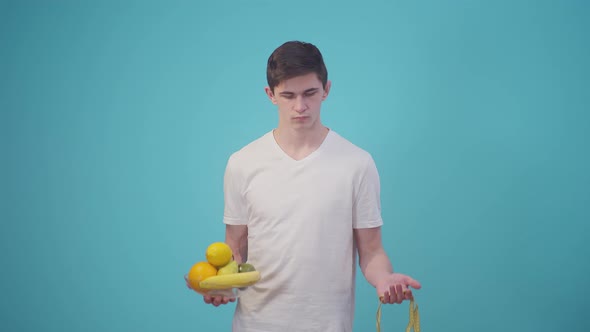 Man in a White Tshirt Holds Fruit and a Meter Ribbon on Blue Background in the Studio alt