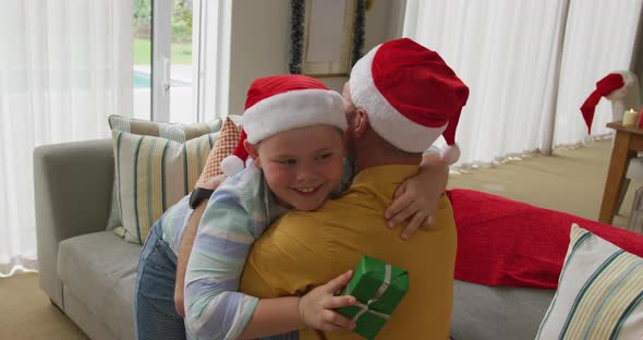 Caucasian boy wearing santa hat holding a christmas gift hugging his father at home alt