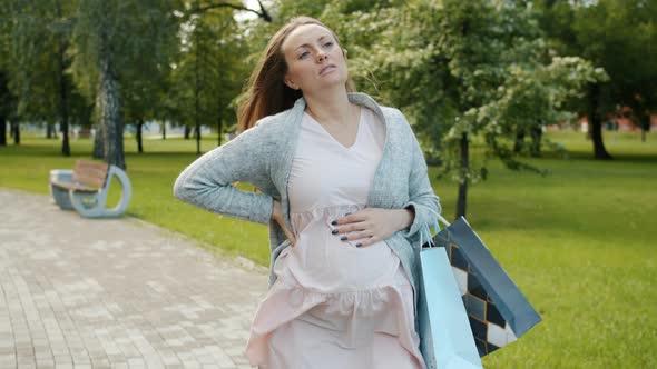 Tired Pregnant Woman Walking in Park Holding Shopping Bags Then Touching Back Suffering From alt