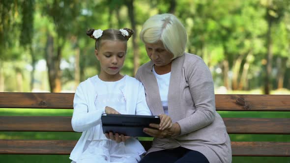 Little Girl and Grandmother Scrolling on Tablet in Park, Shopping App Online alt