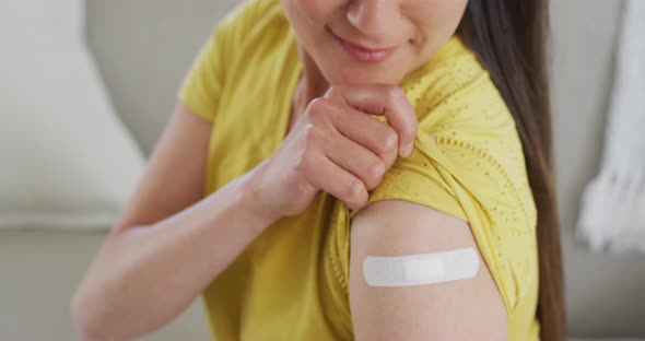Happy asian woman sitting on sofa showing arm with plaster after vaccination alt