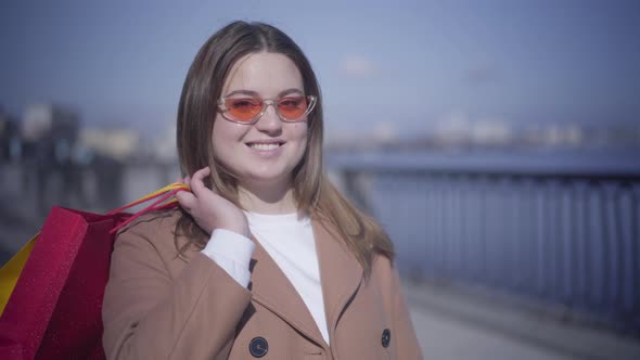 Close-up Portrait of Smiling Caucasian Chubby Girl in Sunglasses Posing with Shopping Bags Outdoors alt