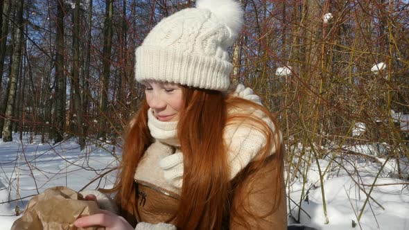 Red-Haired Teen Girl Sitting In Snowy Park And Having A Snack. Girl Laughing