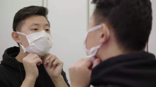 A Young Asian Man in a Face Mask Adjusts the Mask in Front of a Mirror alt