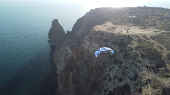 Aerial Drone View of a Man Flying a White and Blue Paraglider Over a Hill and Trees to the Sea Waves alt