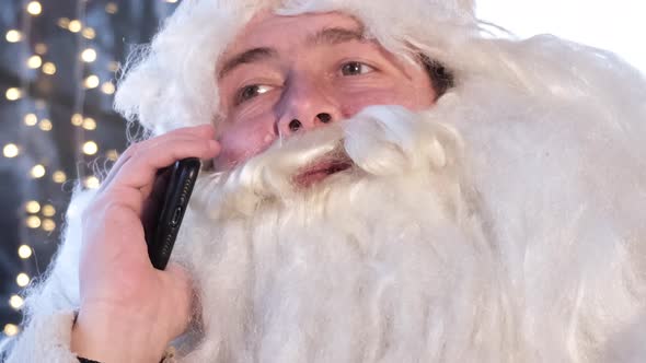 Santa Claus Uses a Smartphone While Sitting in a Cafe He is Resting and Talking on the Phone alt