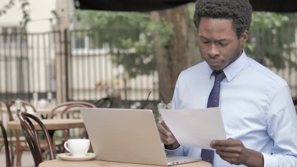 African Businessman Reading Contract and Using Laptop, Outdoor Cafe alt
