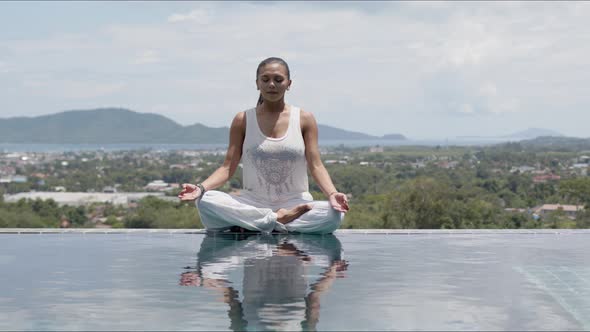 Calm Woman Practicing Yoga in Lotus Posture Poolside Against Resort Town alt