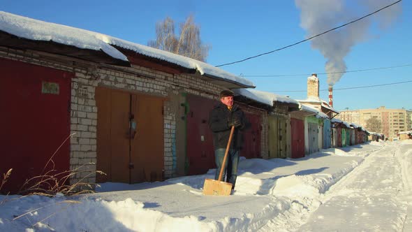 A Man in Warm Clothes is Leaning on a Wooden Shovel Cleansing the Snow alt