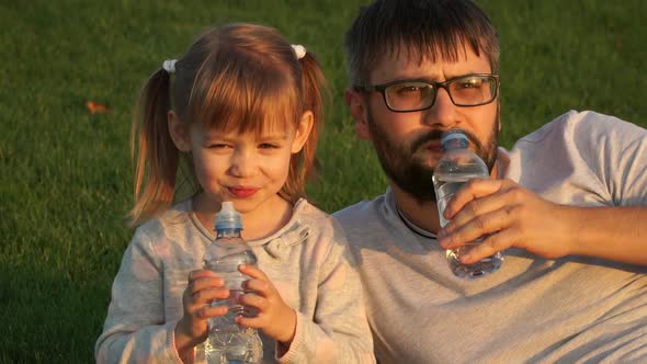 Father with Little Girl Look at Camera Smile Laugh and Drink Bottled Water Outdoors in Summer Park