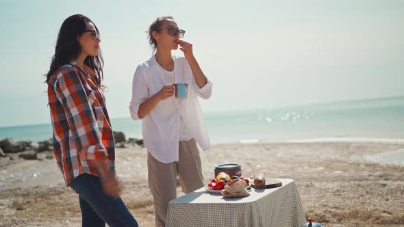 Two Female Friends Stands at Camp Table with Picnic Food on Sea Coast Beach Talking and Eating alt