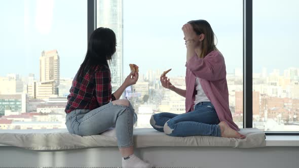 Girls Sit on a Window Sill with a Window and a View of the City Eating Pizza