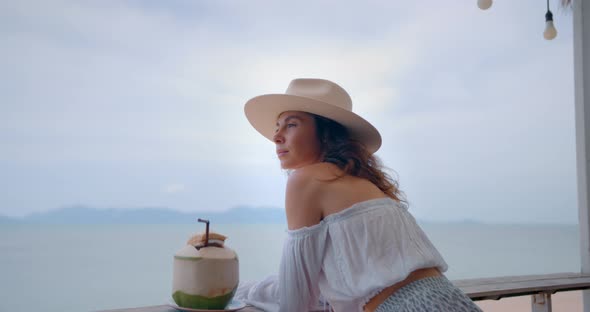 Caucasian Woman in Hat Seats at Cafe Outdoors By the Sea She Drinks Coconut and Enjoys the View alt