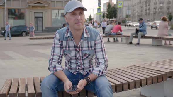 Caucasian Man Sitting on Bench in Square City Holding Smartphone Looking Ahead alt