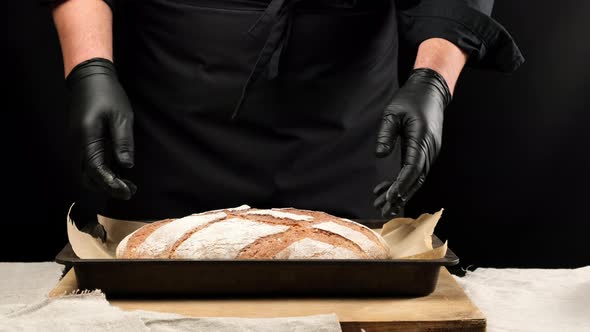 chef in black latex gloves holding baking sheet with baked loaf of rye bread alt