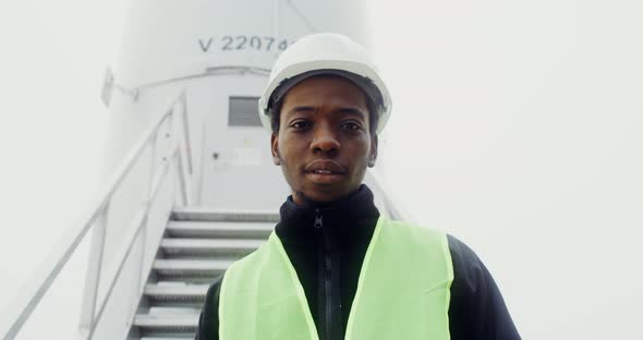 A Man Smiles Looking at the Camera Standing Near an Industrial Wind Turbine alt