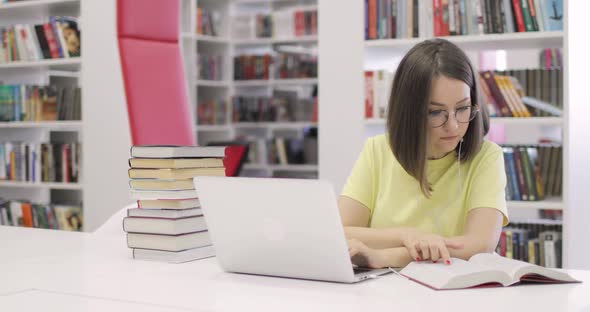 Young Woman is Sitting at a Desk with a Laptop Working in the Library