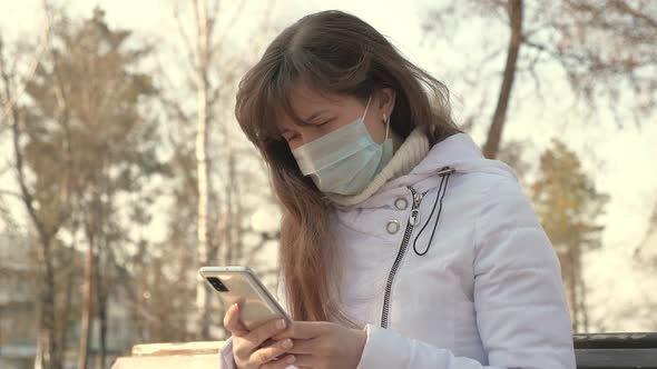 Young Woman in a Medical Protective Mask Is Sitting with a Smartphone on City Street in Europe alt