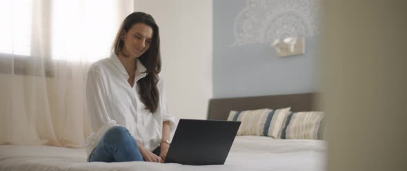A young woman smiling while working on a laptop from her bedroom.  alt