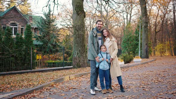 Young Mum Dad and Daughter Schoolgirl with Backpack are Hugging and Smiling Standing in Autumn Park alt