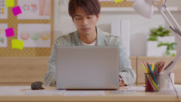 Asian young man sits at his desk and begins turn on and use his laptop for work from his home alt