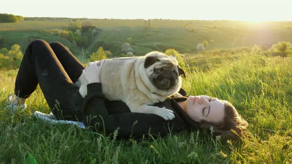 Woman Lies with a Pug in the Grass on Sunset Holds in Her Arms and Petting the Dog