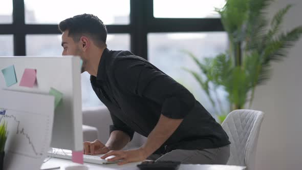 Young Man Typing on Computer Keyboard Standing Up From Table and Walking to Window in Home Office alt