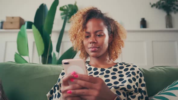 Young African American Woman Chats in Phone Sits on Sofa in Living Room alt