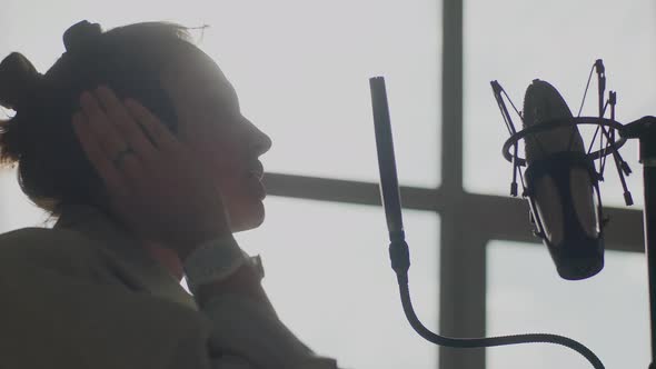A Young Girl Emotionally Sings Into a Microphone in the Background of the Window alt