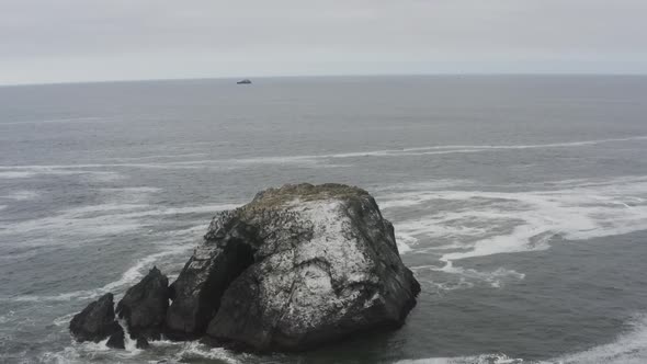 Stormy day aerial of a rocky ocean shore, panning around a large rock in the ocean, waves crash agai alt