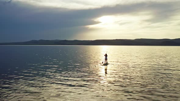 Aerial Shot of a Silhouette of a Woman Floating on a Surfboard with a Paddle on a Mountain Lake at