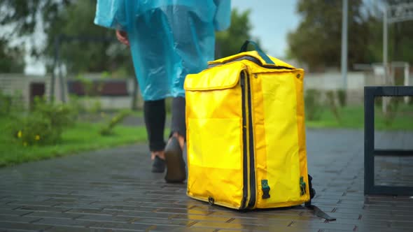 Unrecognizable Young Man in Rain Coat Leaving Yellow Delivery Backpack Walking Away in City Park alt