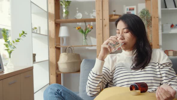 Sick young Asia woman holding pill glass of water take medicine sit on couch at home.