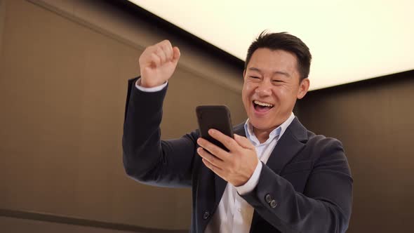 Portrait of Happy Asian Businessman Enjoy Success on Mobile Phone at Office alt