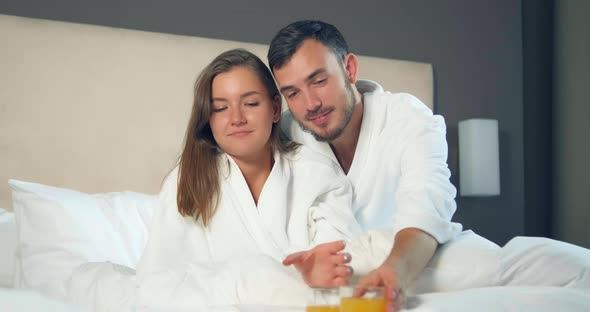 Couple in Bathrobes Drink Orange Juice Lying on Large Bed