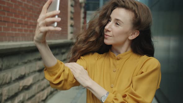 Portrait of Pretty Female Student Taking Selfie Using Smart Phone Camera Against City Background alt