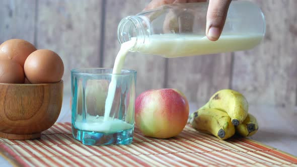 Pouring Milk in a Glass with Banana Egg and Apple on Table alt