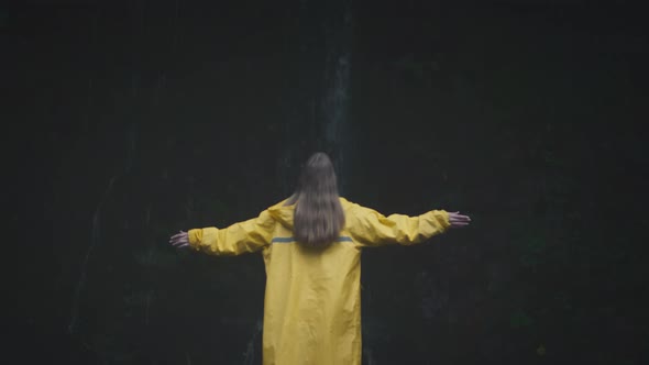 Caucasian Girl Traveler in a Yellow Raincoat Raises Her Hands Near a Waterfall in the Forest alt