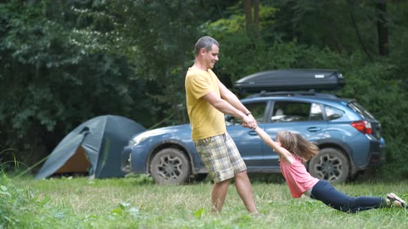 Happy father spinning his smiling daughter child around holding her hands at camping site in summer. alt