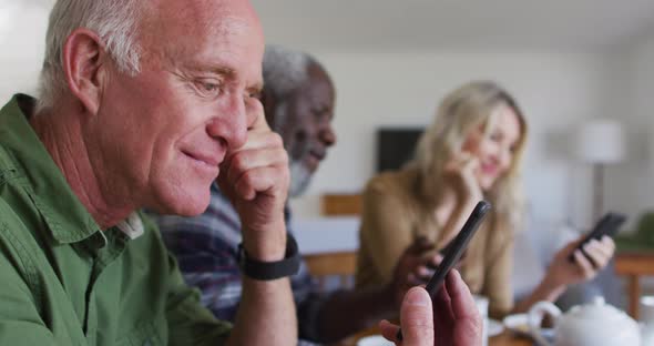 Two diverse senior couples sitting by a table drinking tea using smartphones at home alt
