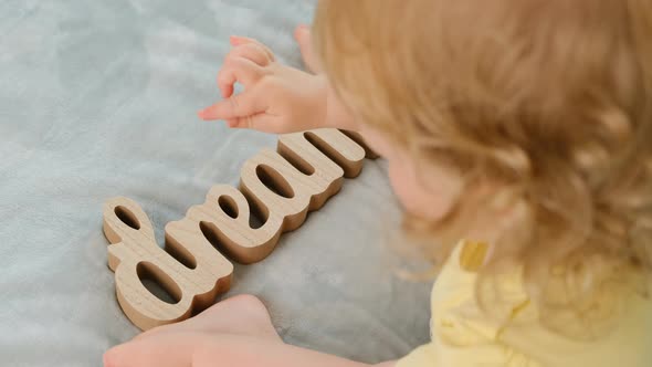 Little Curly Girl Child in Yellow Tshirt Sits on Blue Blanket with Wooden Inscription DREAM Handmade alt