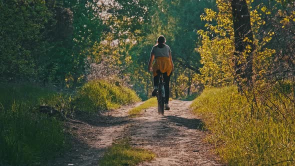 Young Woman on a Bicycle Rides Along Green Forest Path in Sunny Summer Day alt
