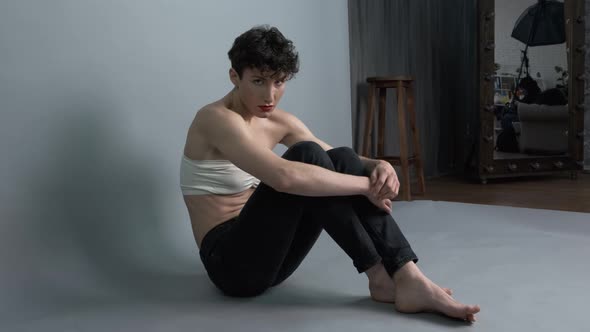 Transgender Guy with Makeup Posing While Sitting on Gray Studio Background alt