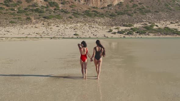 Pretty Female Friends Walking Holding Hands By Sea on Sandy Tropical Beach in Swimsuits alt