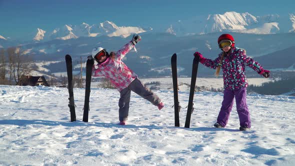 Little Girls in Outwear and Helmets Posing with Skis on Snow with Mountain Range on Background
