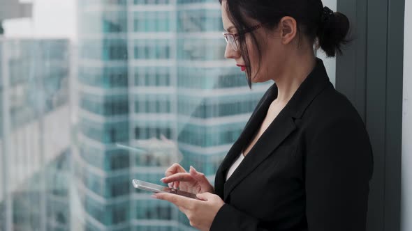 Businesswoman Using a Cell Phone While Standing on a High Floor of a Skyscraper Near a Window. alt