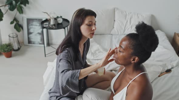 Young Woman Putting Cream on Girlfriends Face alt